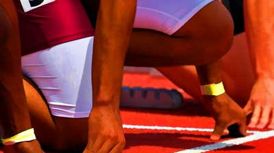 Track athletes in starting position with hands on the red track. One athlete wears a maroon and white uniform with the number 5 visible, preparing for a race. Their figures are partially shown against a blurred background.