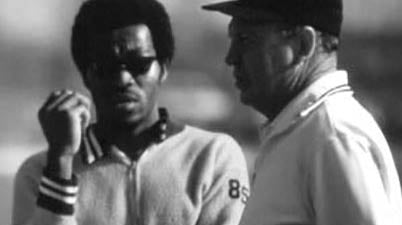 Two men stand outdoors, engaged in conversation. The man on the left wears sunglasses and a jacket, while the man on the right wears a cap and a polo shirt. The image is in black and white.