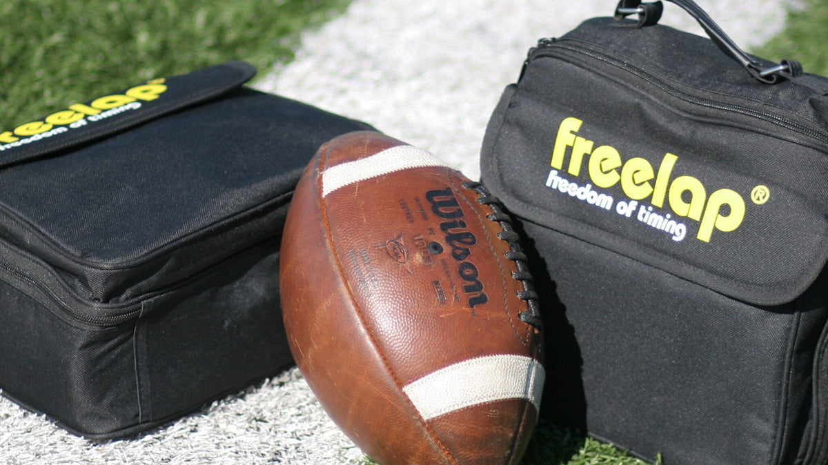 A football, emblematic of sprint-based football action, rests on a white sideline of a grassy field, flanked by two sleek black Freela bags.