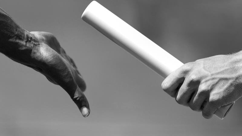 Two hands captured in a close-up as one passes a baton to the other, symbolizing teamwork and exchange within the track team. The background is blurred, focusing attention on the relay handoff in a grayscale setting.