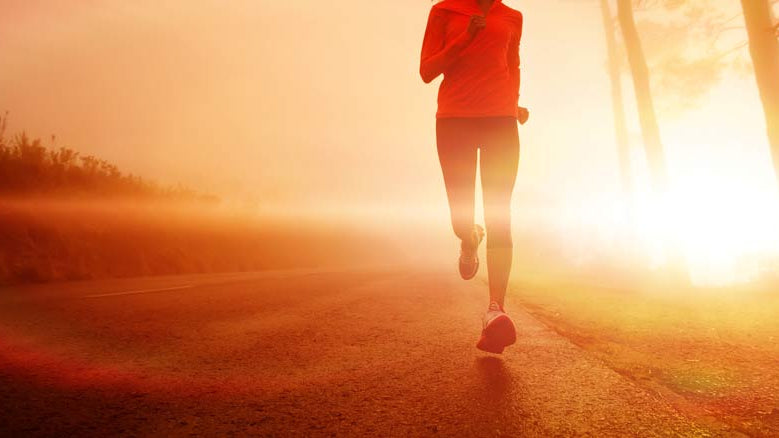 A person jogging on a road during sunrise or sunset, wearing a red jacket. The scene is bathed in warm golden light, with trees visible in the background.
