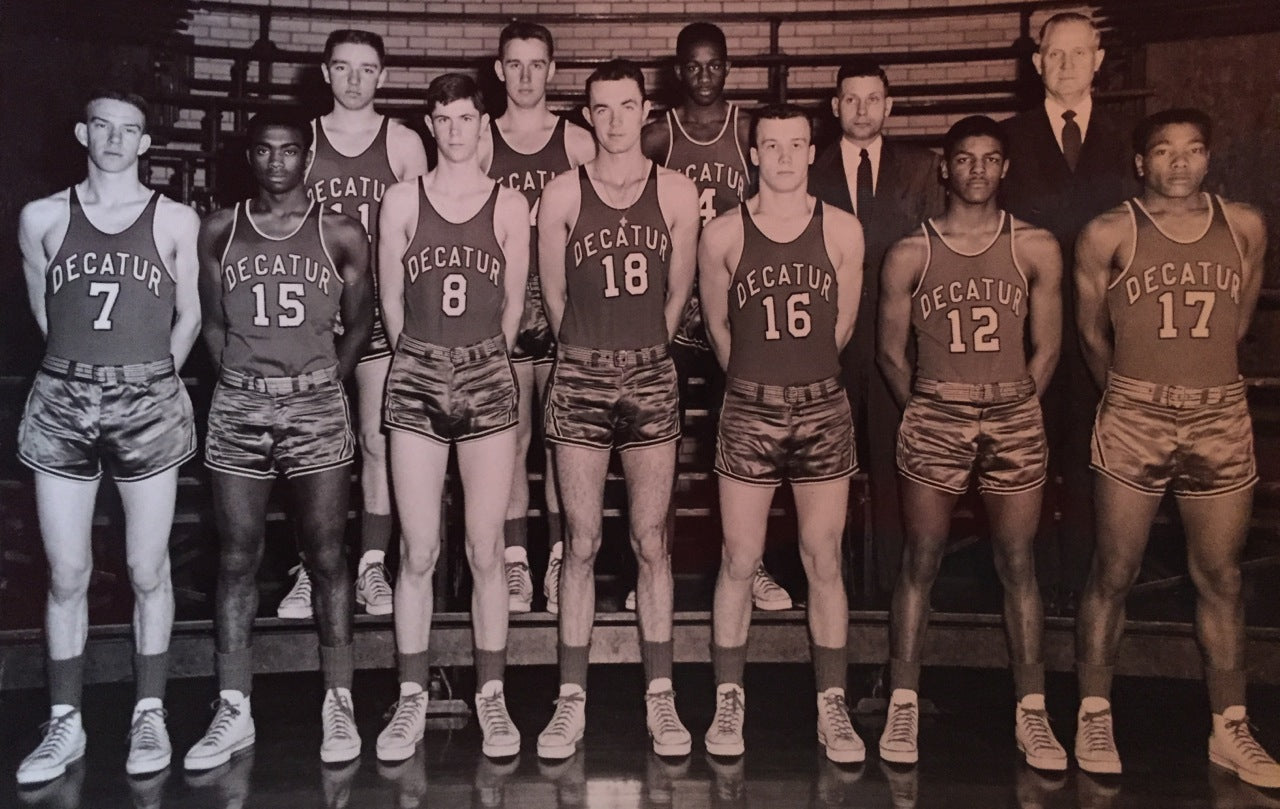 A vintage black and white photo of a basketball team wearing Decatur jerseys. The group consists of eleven players and two coaches. They pose in two rows inside a gym. The players are in uniform with numbers 7, 15, 8, 18, 16, 12, and 17 visible.