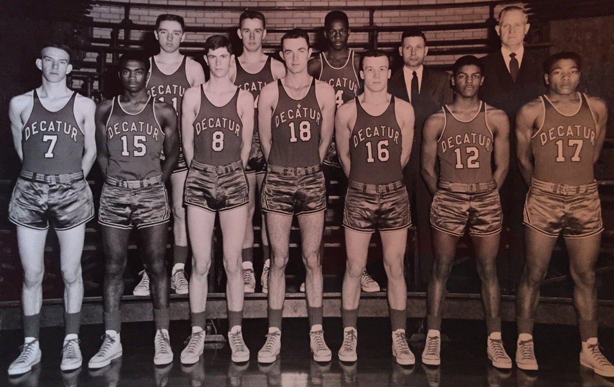 A vintage black and white photo of a basketball team wearing Decatur jerseys. The group consists of eleven players and two coaches. They pose in two rows inside a gym. The players are in uniform with numbers 7, 15, 8, 18, 16, 12, and 17 visible.