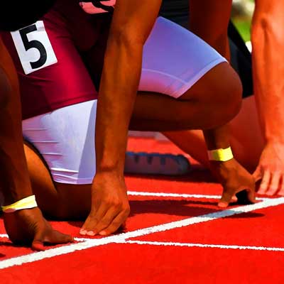 Track athletes in starting position with hands on the red track. One athlete wears a maroon and white uniform with the number 5 visible, preparing for a race. Their figures are partially shown against a blurred background.