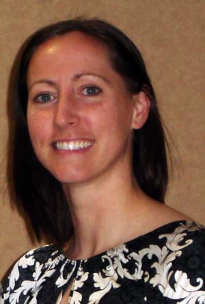 A smiling person with shoulder-length brown hair, wearing a black and white patterned top, stands against a plain beige background.