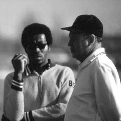 Two men stand outdoors, engaged in conversation. The man on the left wears sunglasses and a jacket, while the man on the right wears a cap and a polo shirt. The image is in black and white.