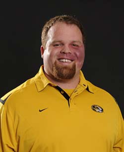 A person with a beard and short hair is smiling while wearing a yellow collared shirt with a logo. The background is a solid dark color.
