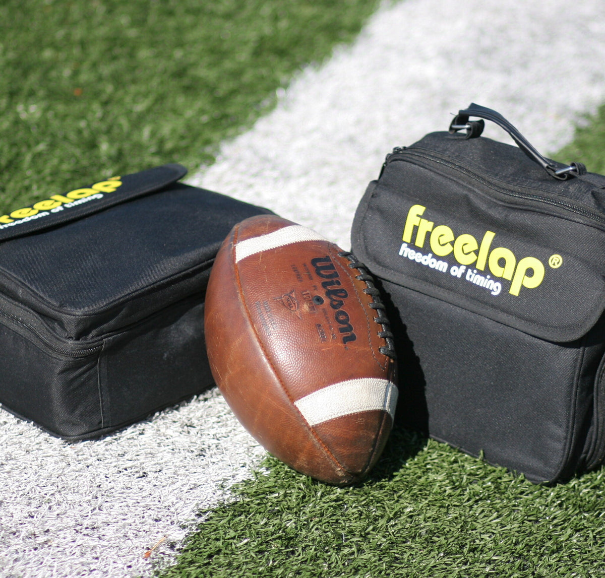 A football, emblematic of sprint-based football action, rests on a white sideline of a grassy field, flanked by two sleek black Freela bags.