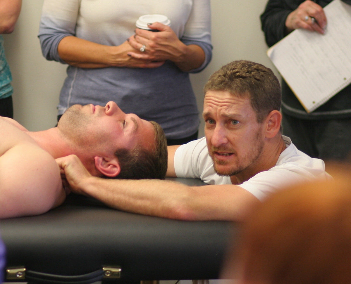 A person lying on a massage table while another individual demonstrates a neck massage technique. Several people are observing, some holding notes and a cup. The setting appears educational or instructional.