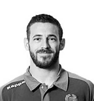 A man with short dark hair and a beard is wearing a collared shirt with the Kappa logo. The image is in black and white.