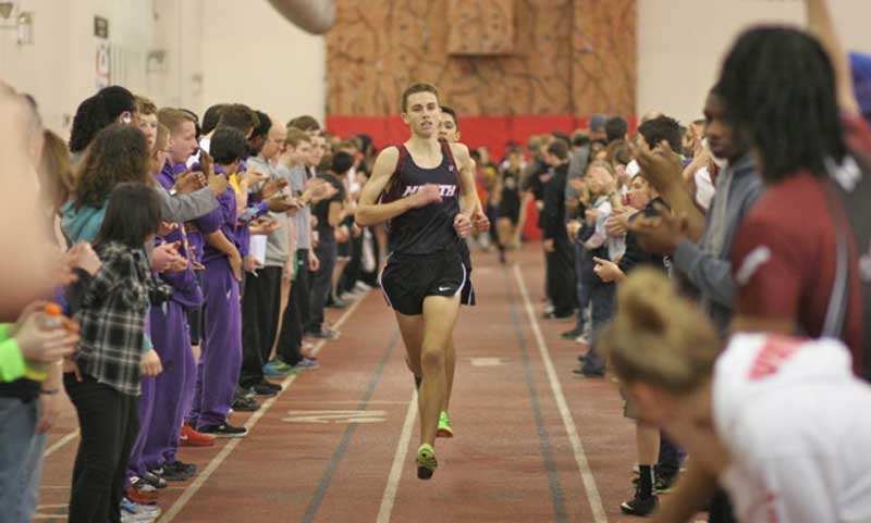 A runner in a purple singlet sprints down an indoor track while a crowd of people lines both sides, cheering and clapping. The background features a large red and brown climbing wall.