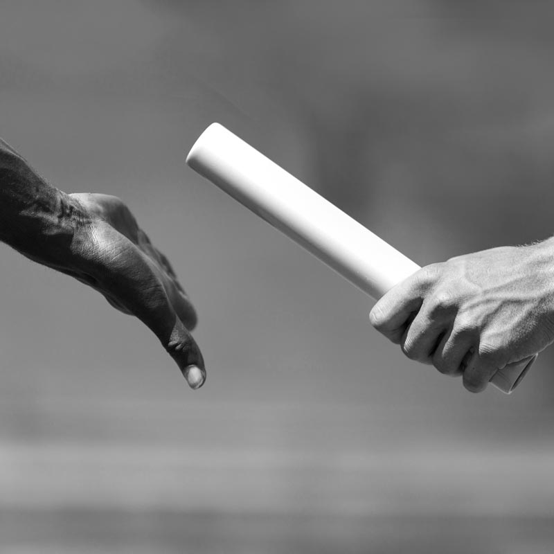 Two hands captured in a close-up as one passes a baton to the other, symbolizing teamwork and exchange within the track team. The background is blurred, focusing attention on the relay handoff in a grayscale setting.