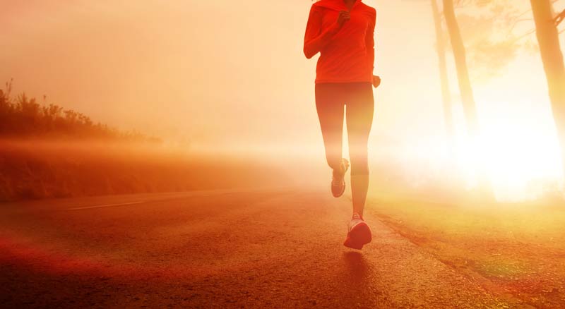 A person jogging on a road during sunrise or sunset, wearing a red jacket. The scene is bathed in warm golden light, with trees visible in the background.