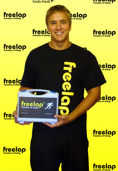 A person, Alex Taray, smiles while holding a white box with freelap branding. They wear a black shirt showcasing the same logo. The yellow background is adorned with repeated freelap logos.