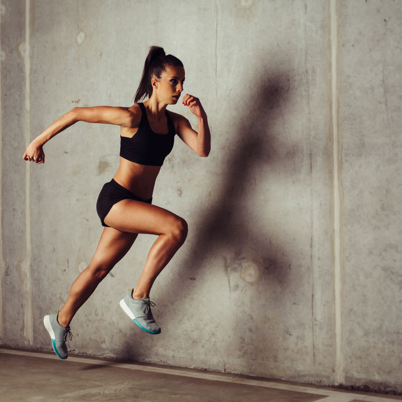 A person in athletic wear leaps in mid-air against a concrete wall backdrop. The individual has their hair in a ponytail and is wearing a black sports bra, black shorts, and white sneakers, showcasing energy and strength.